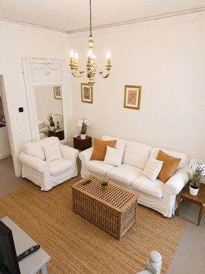 Een woonkamer met een witte zithoek, een houten salontafel, een kroonluchter en kunstwerken in lijsten aan de wanden.