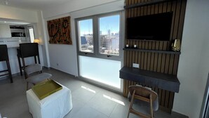 A modern living room with a large window, a flat-screen TV mounted on a wooden panel, and a small kitchen area in the background. - Fantastic Studio With Balcony Pool (Buenos Aires)