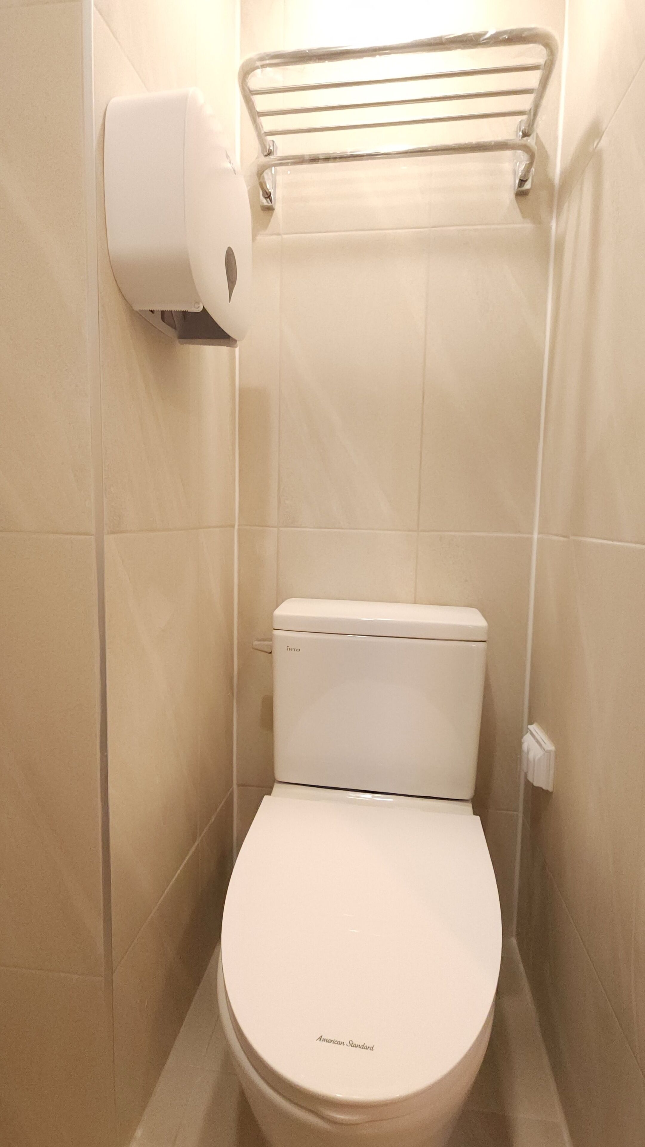 A small, white toilet with a 'hro' label, a paper towel dispenser, and a metal towel rack in a tiled bathroom.