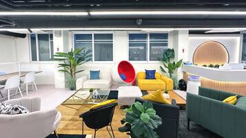 Un espacio de oficina moderno con una variedad de zonas de estar, incluyendo un sofá blanco, un sofá amarillo y un sofá verde. Hay varias plantas, una mesa de centro de vidrio y una silla roja. La habitación tiene ventanas amplias y un mostrador de recepción blanco con un panel de madera circular.