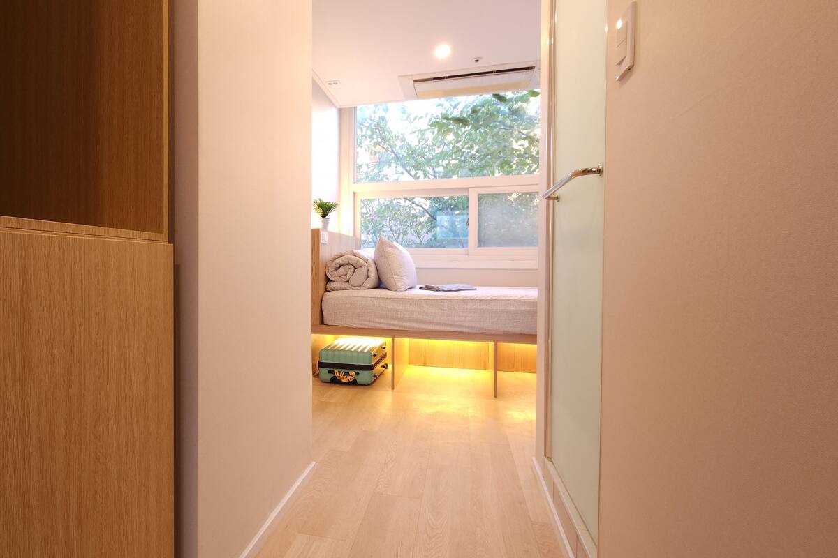 Een smalle gang die uitkomt in een kamer met een bed, een raam en een koffer op de vloer.