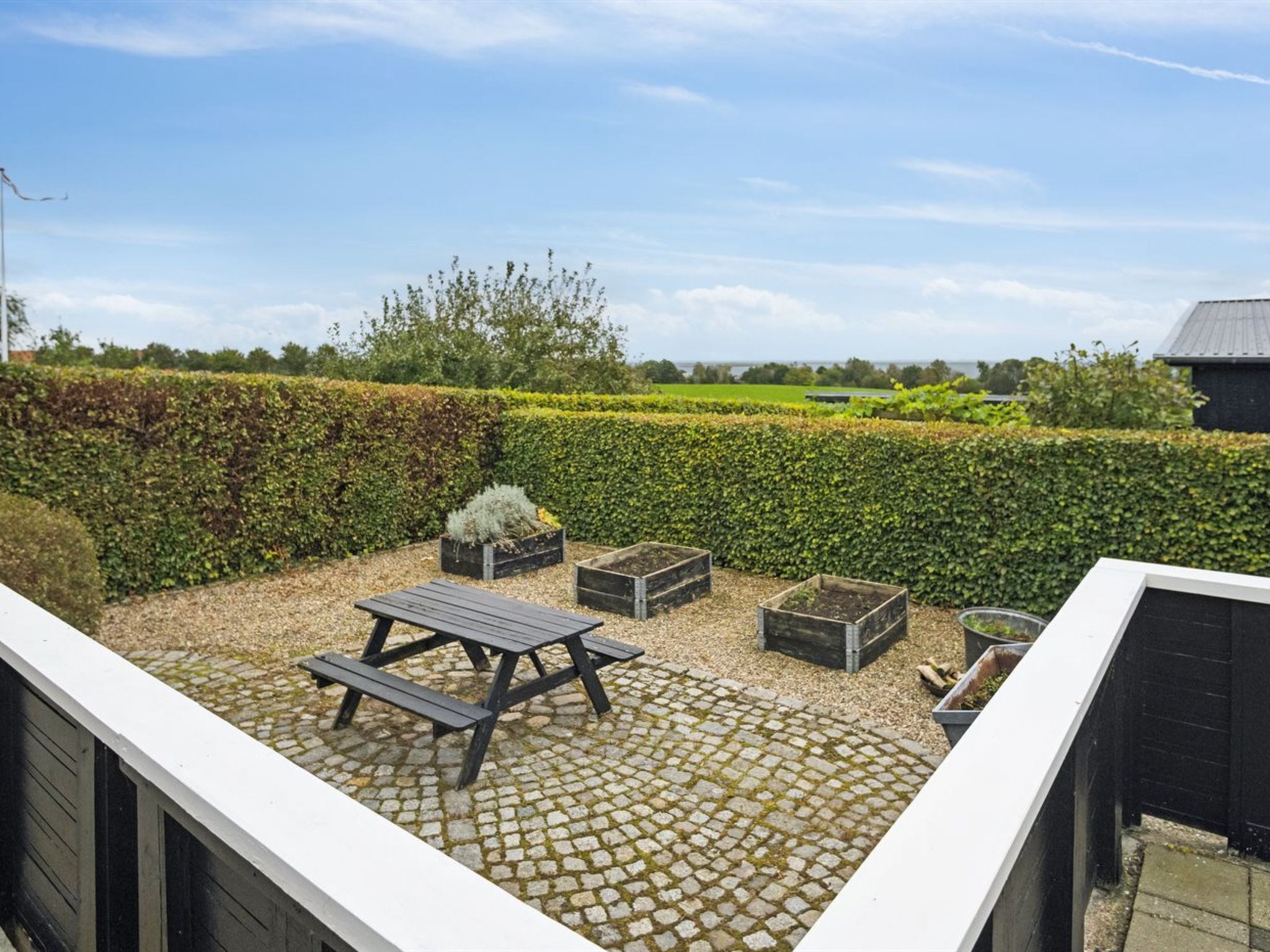 Une terrasse avec une table et des bancs en bois, entourée d’une haie et offrant une vue sur la campagne.