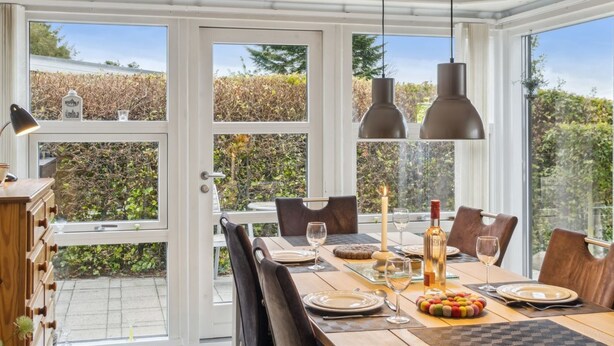 Un comedor con una mesa dispuesta para cuatro, ventanas amplias y una estantería sobre ellas.