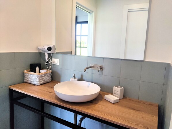 Un baño con tocador de madera, un lavabo blanco, un secador de pelo y una cesta con artículos de aseo.