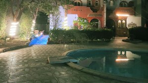 A nighttime view of a hotel courtyard with a swimming pool, a well-lit pathway, and a building with a red facade.
