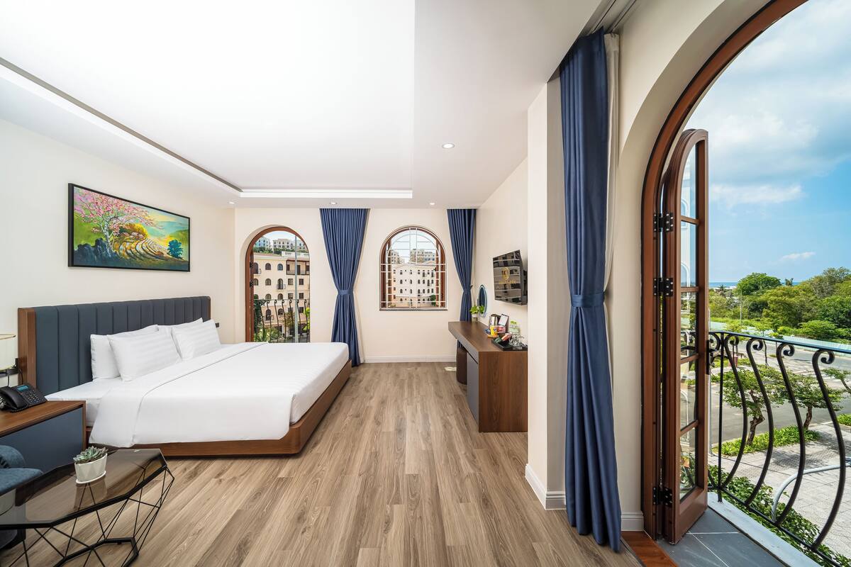 A modern hotel room with a large bed, a desk, and a view of the city through arched windows.