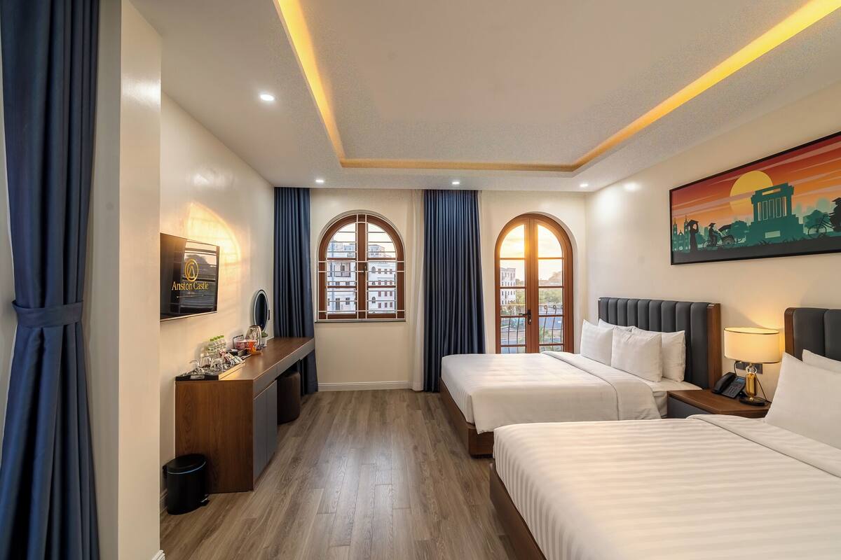 A hotel room with two beds, a desk with a computer, a window with curtains, and a painting on the wall.