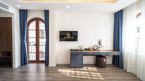 A hotel room with a wooden desk, a television mounted on the wall, a large window with curtains, and a wooden floor. - Anston Castle Hotel (Phu Quoc)