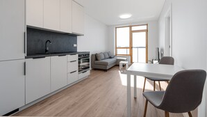 A modern kitchen with white cabinets, a black countertop, and a built-in oven. There is a gray sofa and a small table with a chair in the living area. - Cozy two room apartment near city center (Vilnius)