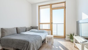 A modern bedroom with a large bed, a small white table, a TV, and a large window with a view. - Cozy two room apartment near city center (Vilnius)