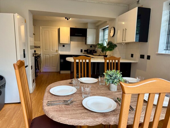 A dining area with a round table set for four, surrounded by wooden chairs. There is a potted plant on the table, and the room has a modern kitchen in the background.