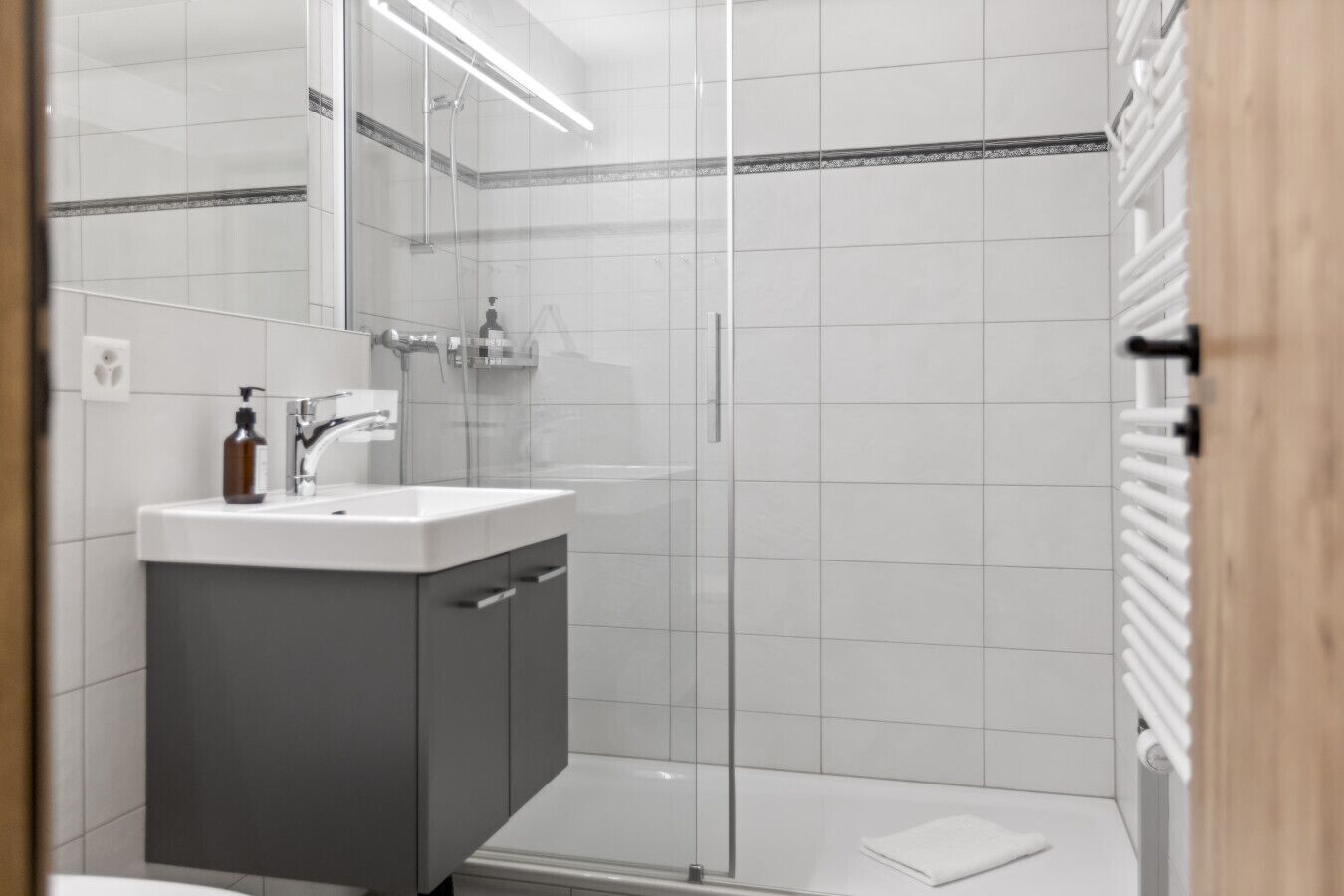 Ein modernes Badezimmer mit Duschkabine aus Glas, einem weißen Waschbecken inklusive Spiegel und einem Handtuchhalter.