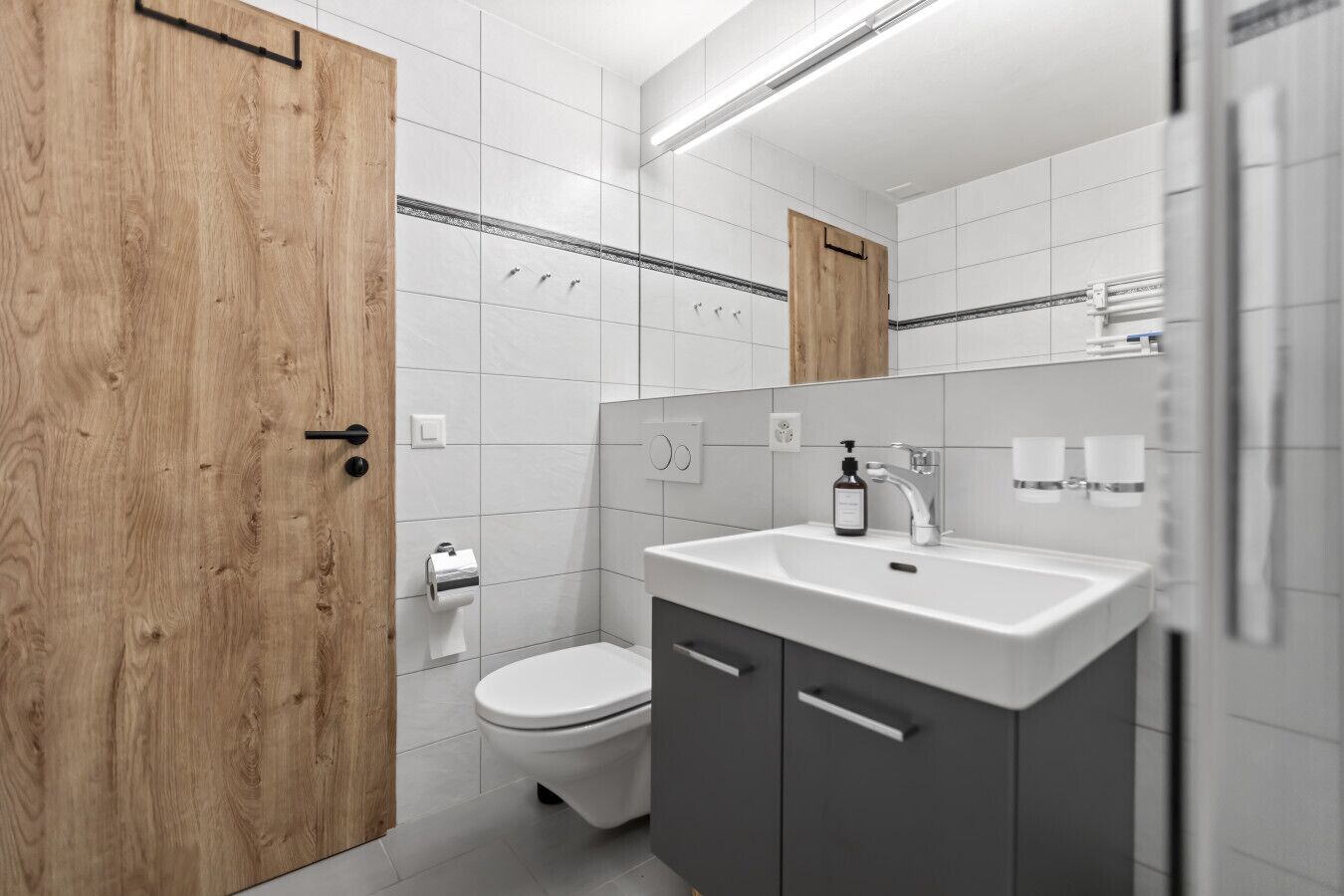 Un baño moderno con puerta de madera, azulejos blancos, inodoro, lavamanos con espejo y dispensador de toallas.
