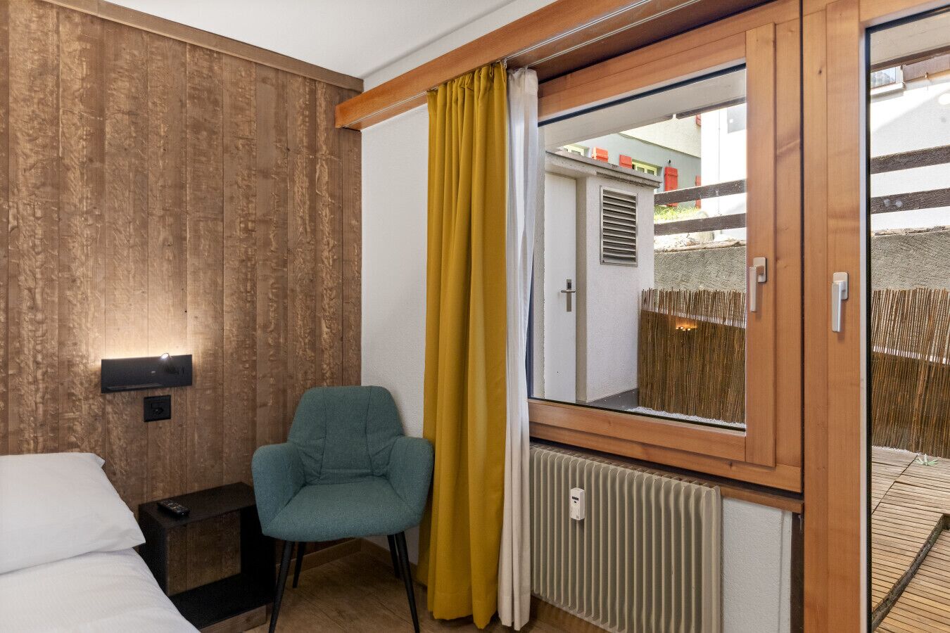 Ein Hotelzimmer mit einer Holzwand, einem Einzelbett, einem türkisblauen Sessel, einem Fenster mit gelbem Vorhang und Blick auf ein Gebäude.