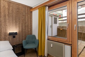 A hotel room with a wooden wall, a single bed, a teal chair, a window with a yellow curtain, and a view of a building outside. - Saas-Fee center - 2 bedrooms - balcony and patio (Saas-Fee)