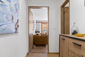 Ein schmaler Flur mit einer Holztür, die in einen Wohnbereich führt, einer Küchenzeile mit einer Glasflasche und einer gelben Schüssel.