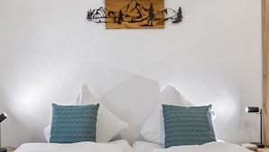 A neatly made bed with white bedding and two teal pillows, a wooden wall art depicting a mountain scene, and a bedside lamp on each side. - Saas-Fee center - 2 bedrooms - balcony and patio (Saas-Fee)