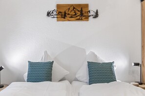 A neatly made bed with white bedding and two teal pillows, a wooden wall art depicting a mountain scene, and a bedside lamp on each side. - Saas-Fee center - 2 bedrooms - balcony and patio (Saas-Fee)