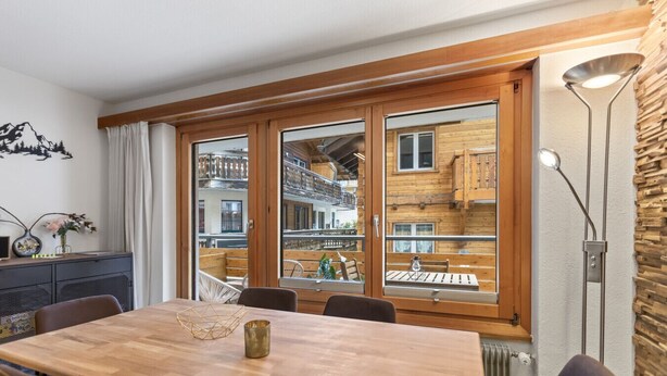 A dining area with a wooden table, chairs, and a view of a building with balconies. - Saas-Fee center - 2 bedrooms - balcony and patio (Saas-Fee)