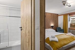 Ein Hotelzimmer mit Holztür, einem Bett mit gelber Decke, einem blauen Sessel und einer Duschkabine.