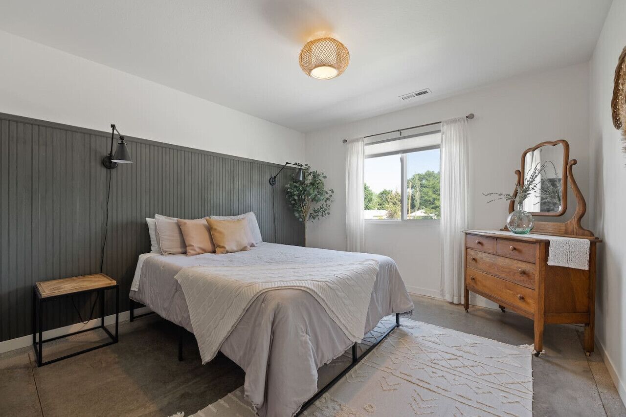 Un dormitorio con cama, mesita de noche, cómoda, espejo y ventana con cortinas.