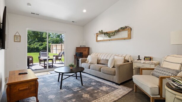 Amplia sala de estar con un sofá grande, una mesa de centro y un televisor montado en la pared. Cuenta con una puerta corrediza de vidrio que da a un patio con mobiliario exterior.