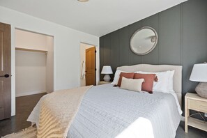 Ein Schlafzimmer mit einem großen Bett, einem Nachttisch mit Lampe, einem Spiegel an der Wand und einer Tür zu einem anderen Raum.