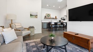 Una sala de estar moderna con un sofá, una mesa de centro y un televisor. Se ve una cocina al fondo con una mesa de comedor y sillas.