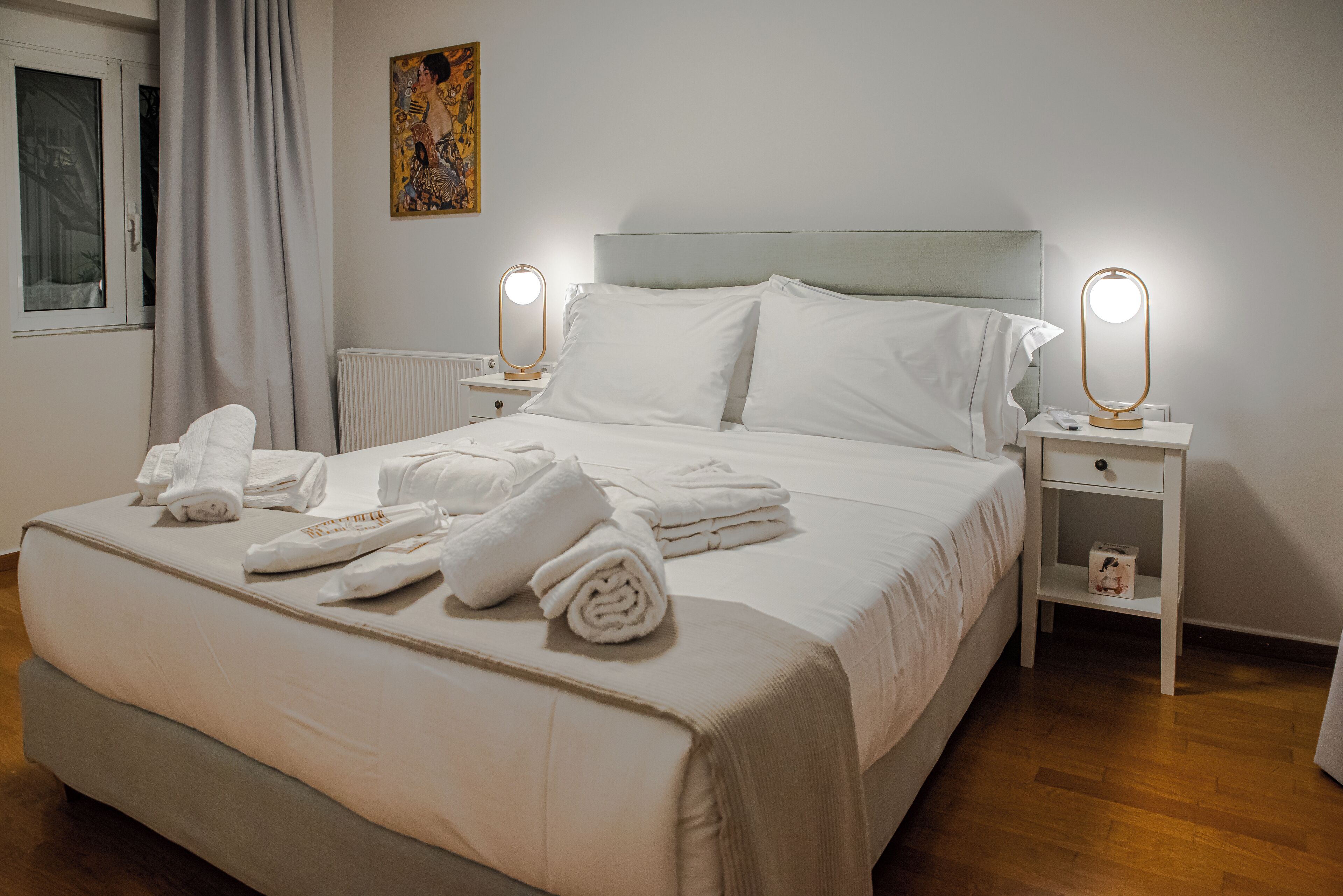 Cama bem arrumada com lençóis e toalhas brancos, cabeceira, mesas de cabeceira com abajures e uma janela com cortinas.