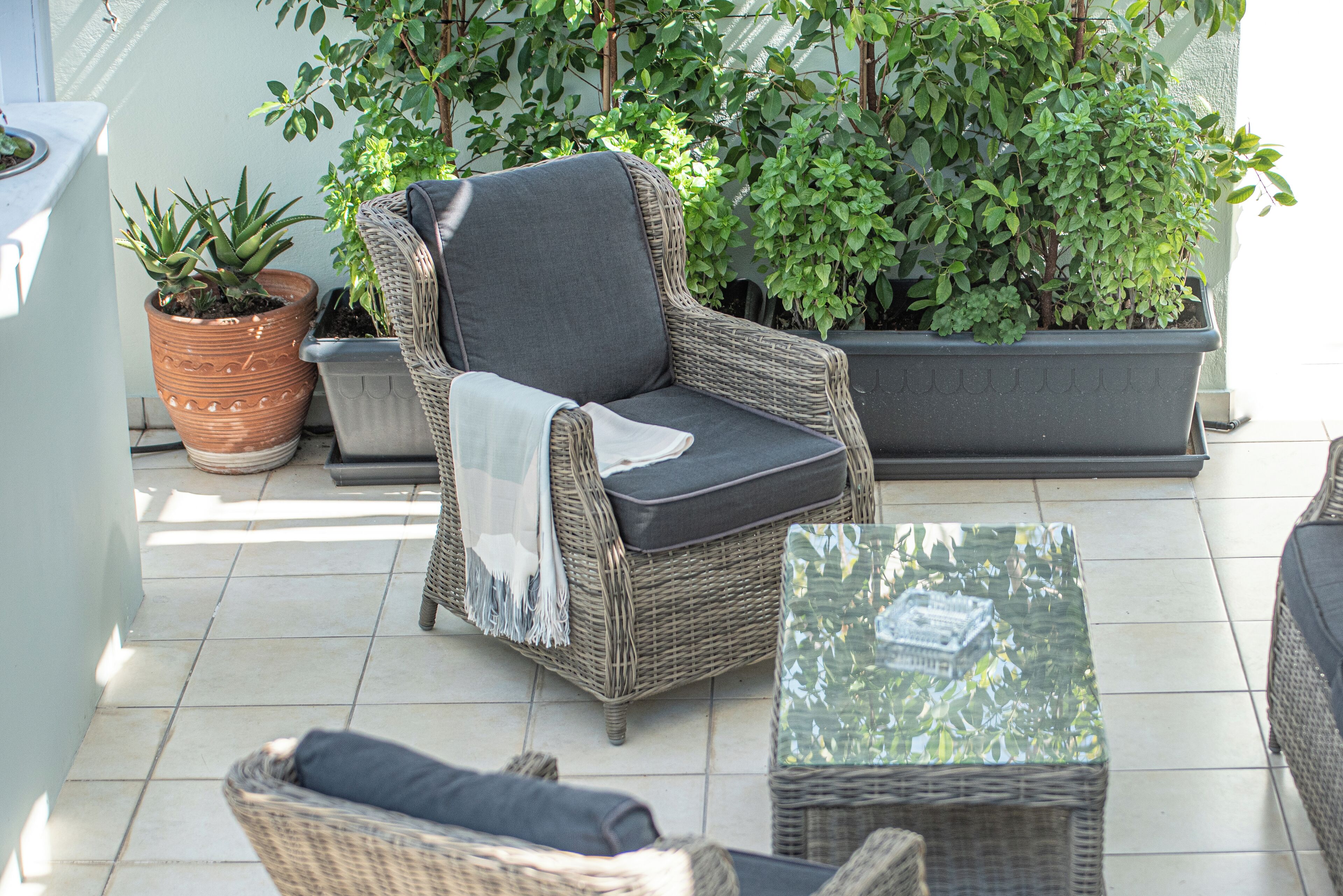 A wicker chair with a cushion, a glass-top table, and potted plants in a sunlit outdoor area.