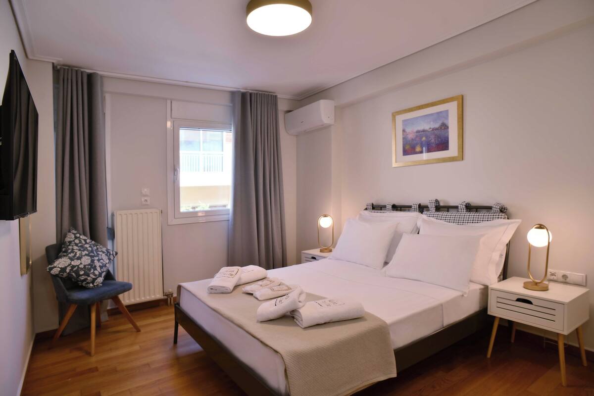 Une chambre d’hôtel bien rangée, avec un lit, des tables de chevet, une chaise, une fenêtre avec des rideaux, un appareil de climatisation et un tableau encadré au mur.
