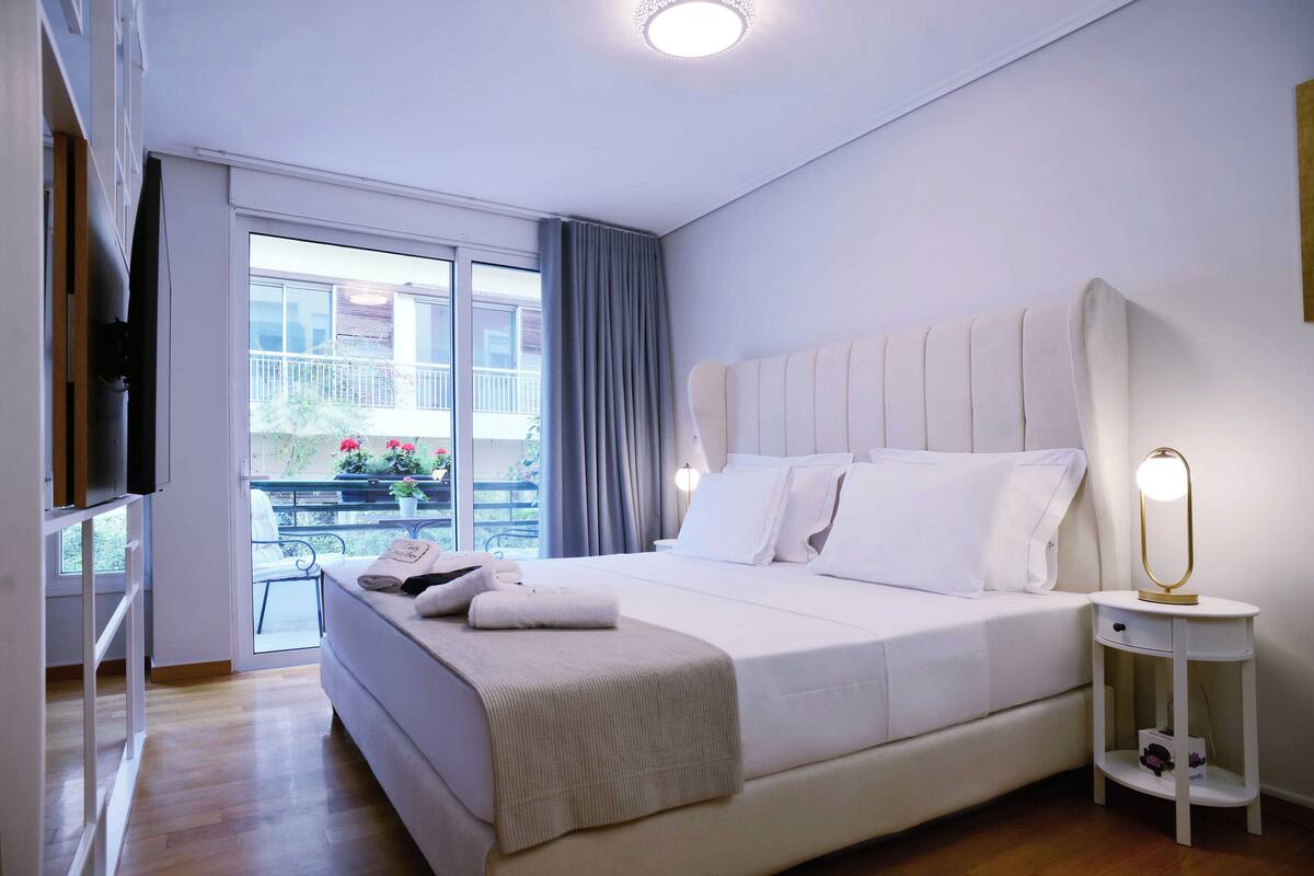 Une chambre d’hôtel moderne avec un grand lit, une table de chevet avec une lampe, un balcon agrémenté de plantes et une porte-fenêtre coulissante.