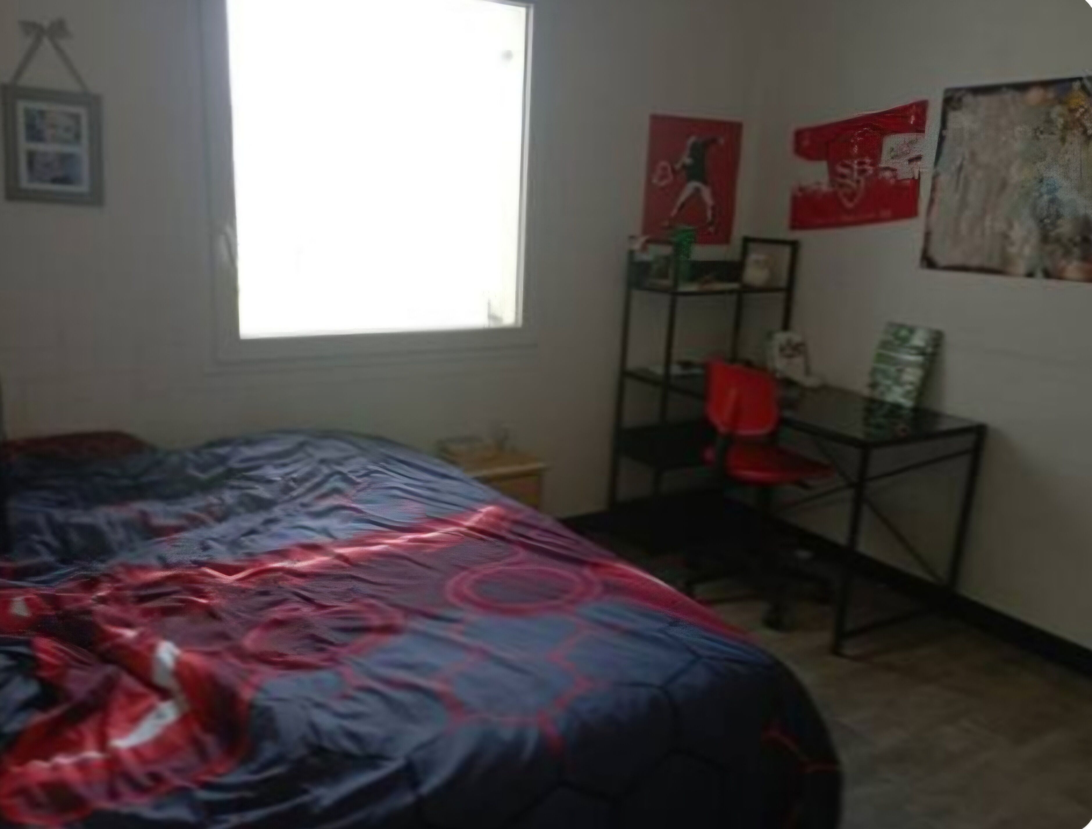 A bedroom with a bed, a desk, and a chair. There is a window providing natural light.