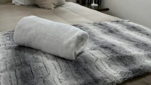 Una cama bien hecha con una almohada blanca y una toalla blanca enrollada sobre una manta gris.