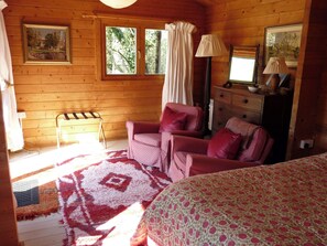 Un dormitorio de cabaña de madera con una cama, dos sillas, un aparador y una ventana con cortinas.