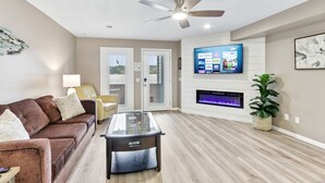 A living room with a brown leather sofa, a glass coffee table, a ceiling fan, a flat-screen TV mounted on a white wall, and a fireplace. - Sandy Toes at Lighthouse Cove (Wisconsin Dells)