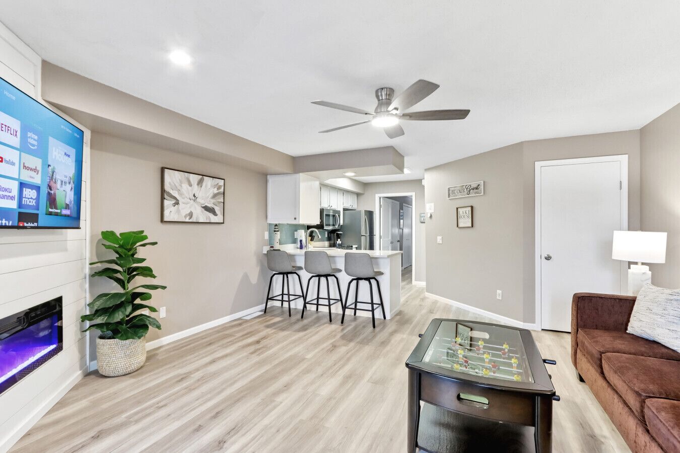 A modern living room with a flat-screen TV, a ceiling fan, a coffee table, a sofa, and a kitchen area with bar stools.