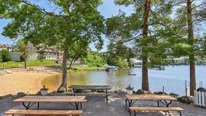 A lakeside patio with picnic tables, a boat on the water, and a building in the background. - Sandy Toes at Lighthouse Cove (Wisconsin Dells)