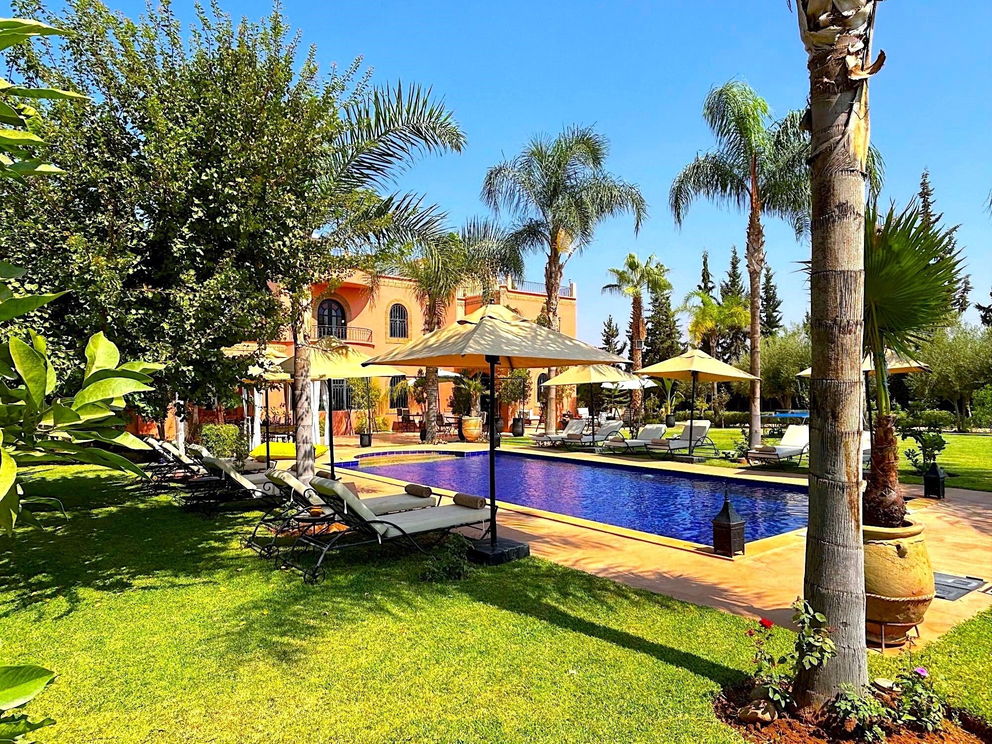 A pool area with lounge chairs and umbrellas, surrounded by palm trees and a building with arched windows.