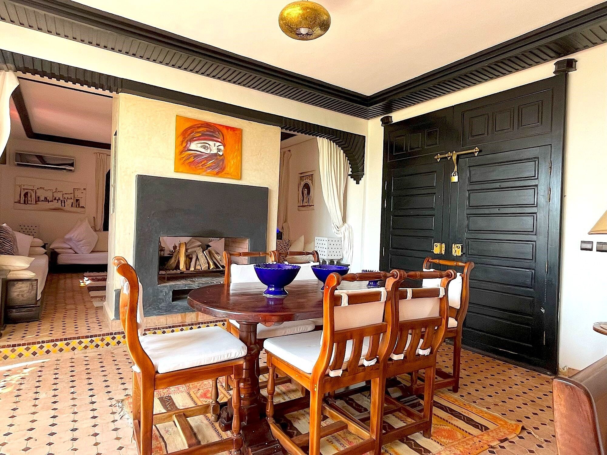 Un coin repas avec une table ronde en bois et quatre chaises, une porte noire, une cheminée et un tableau coloré accroché au mur.