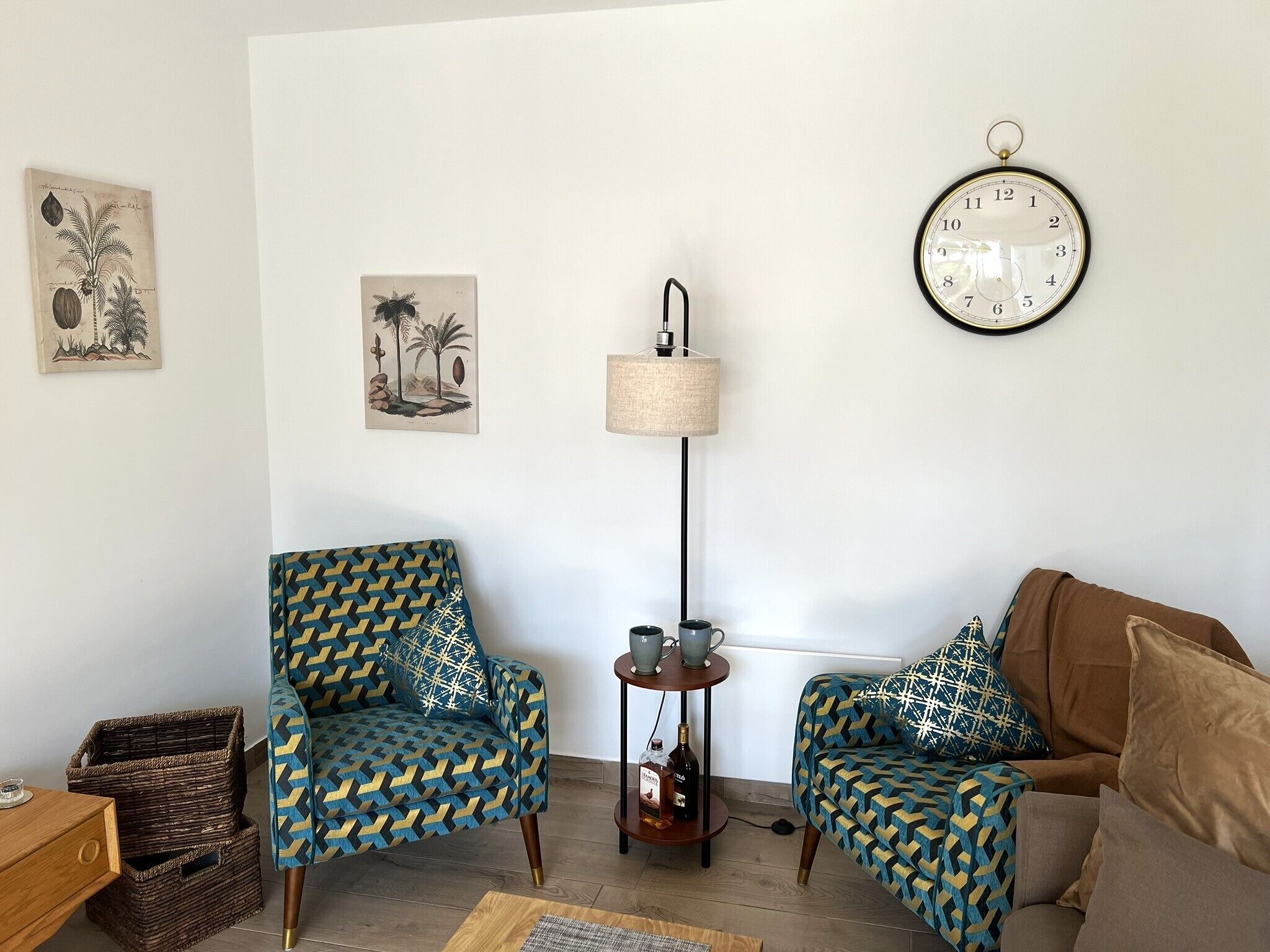 Een woonkamer met twee gestoffeerde fauteuils, een klein bijzettafeltje met een lamp, een wandklok en twee ingekaderde botanische prints.