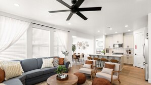 A modern living room with a gray sofa, wooden coffee table, and brown ottomans. There is a dining area with a white table and chairs, and a kitchen with white cabinets and a black ceiling fan. - Escape to your own private oasis in sunny Washington, Utah! (Washington)
