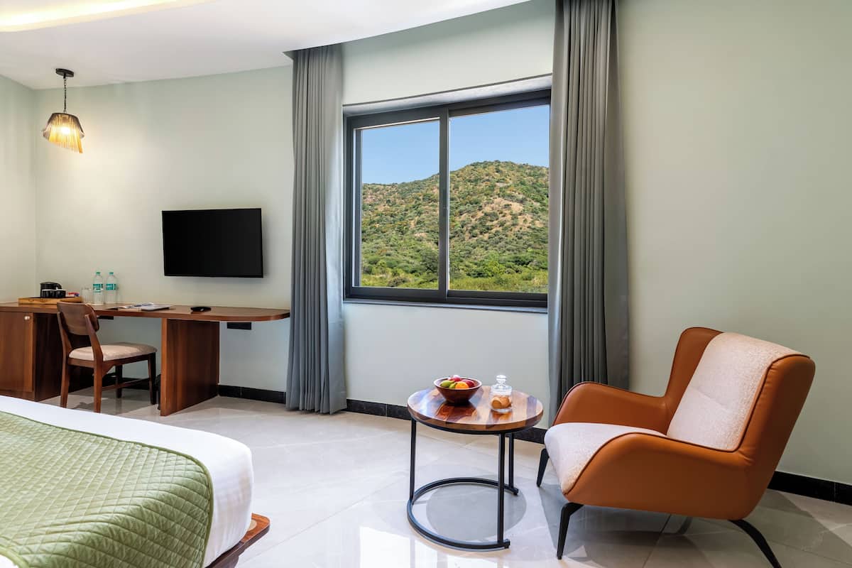 A hotel room with a bed, a desk, a chair, a small table with a bowl of fruit, a bottle, and a TV on the wall.