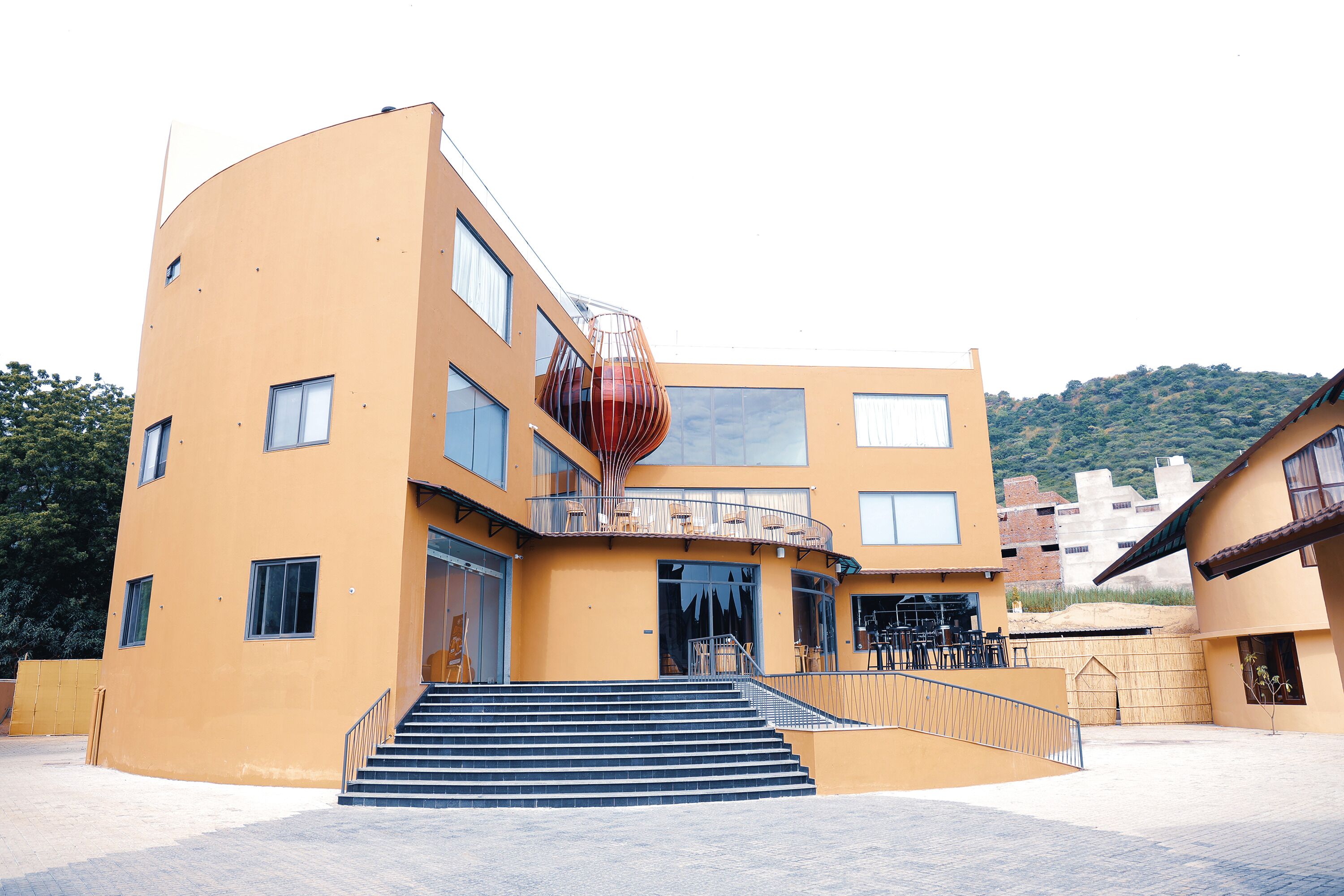 A modern building with a curved facade, a staircase leading to a balcony, and a view of a hilly landscape in the background.