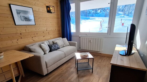 Ein gemütliches Zimmer mit Holzboden, einer Couch mit Kissen, einem kleinen Tisch, einem Fernseher auf einem Schrank und einem Fenster mit Blick auf eine verschneite Landschaft.