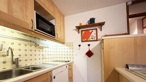 A compact kitchen with wooden cabinets, a stainless steel sink, a microwave, and a small shelf with a kettle and a framed picture. - Spacious 2-room apartment for 6, near shops and slopes (Les Orres)