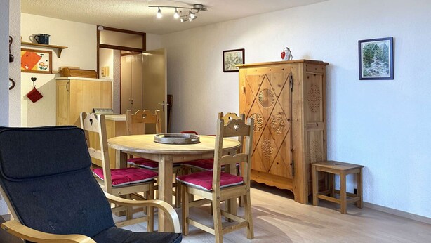A dining area with a round wooden table, four wooden chairs with red cushions, a blue upholstered chair, and a wooden cabinet with intricate carvings. - Spacious 2-room apartment for 6, near shops and slopes (Les Orres)