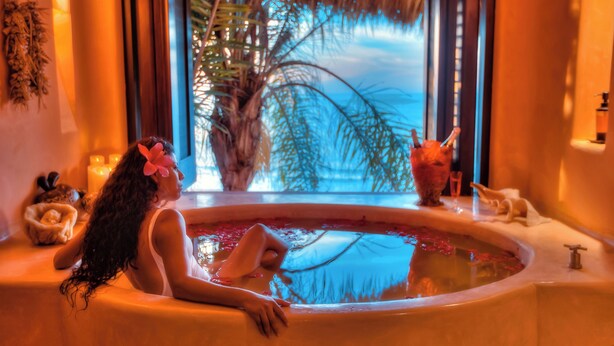 A woman relaxing in a bathtub with a view of a palm tree and a thatched roof structure outside.