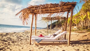 A beach hut with a thatched roof, a white lounge chair, and a red hat on the sand.