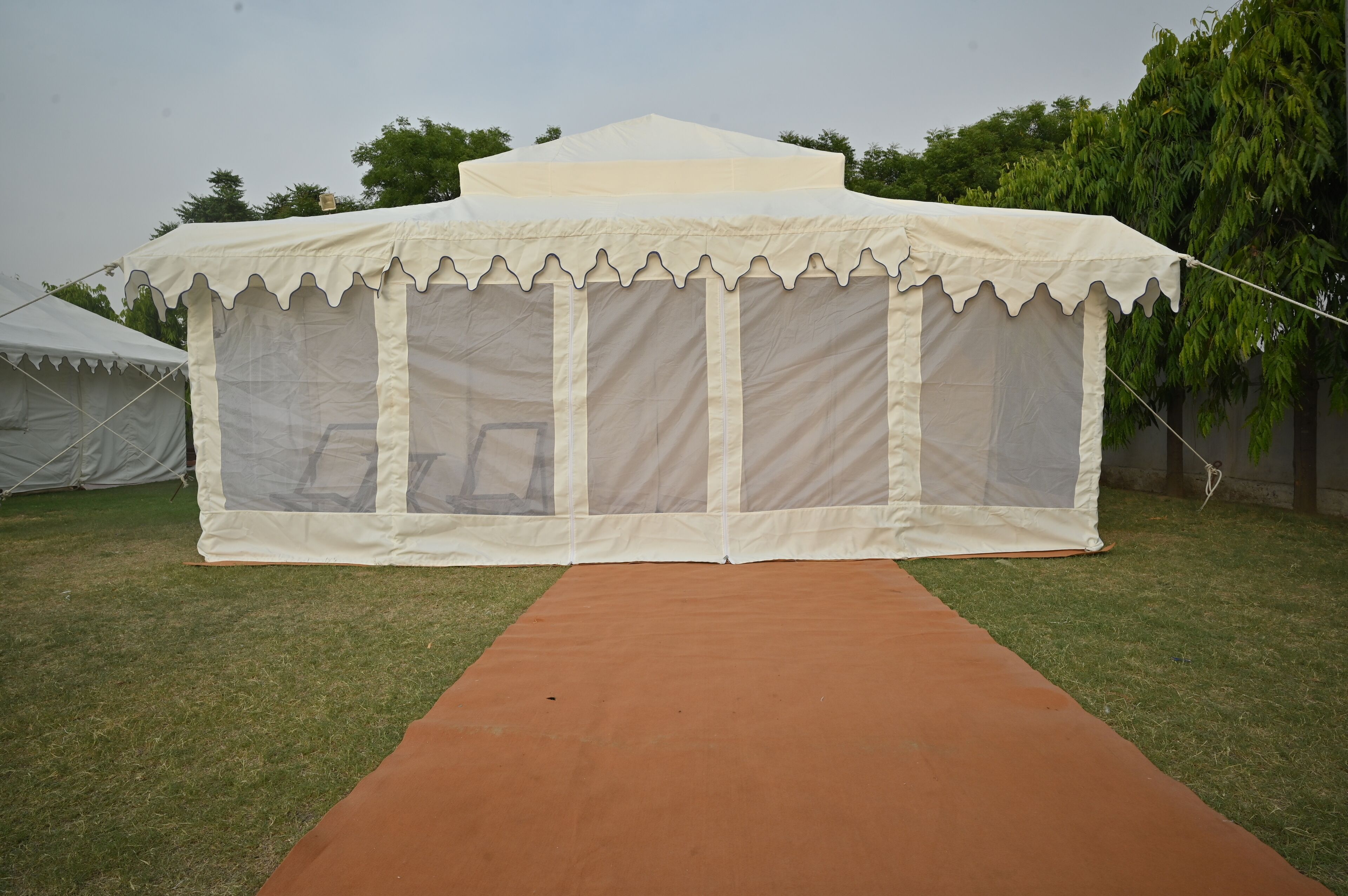 Una carpa grande de exterior blanco y interior marrón, montada en un campo cubierto de césped, con una alfombra roja que la conduce.